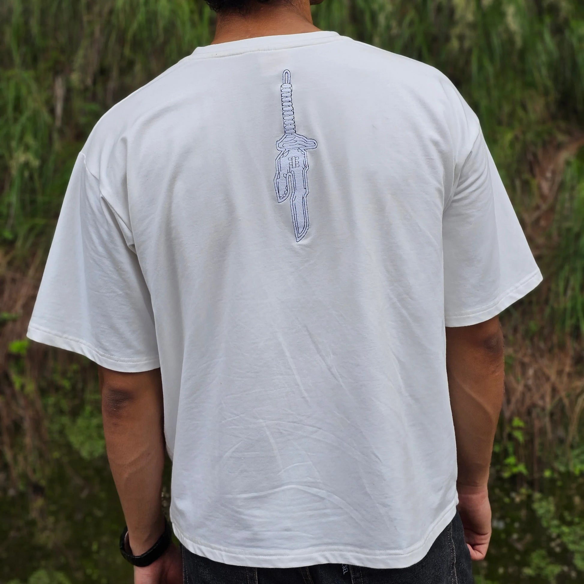 Person wearing a white t-shirt with a sword design on the back, standing outdoors.