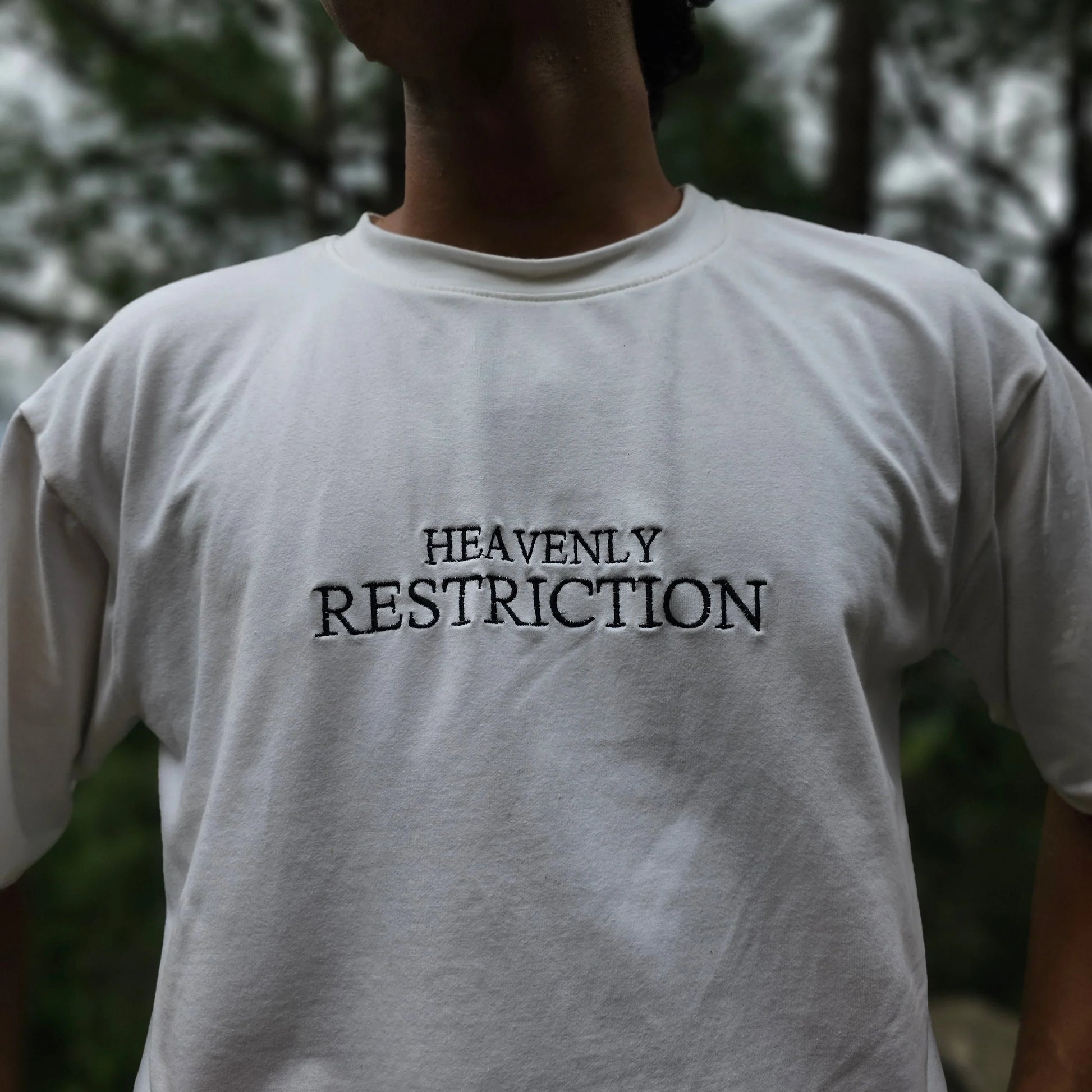 Person wearing a white shirt with 'HEAVENLY RESTRICTION' text, standing outdoors.