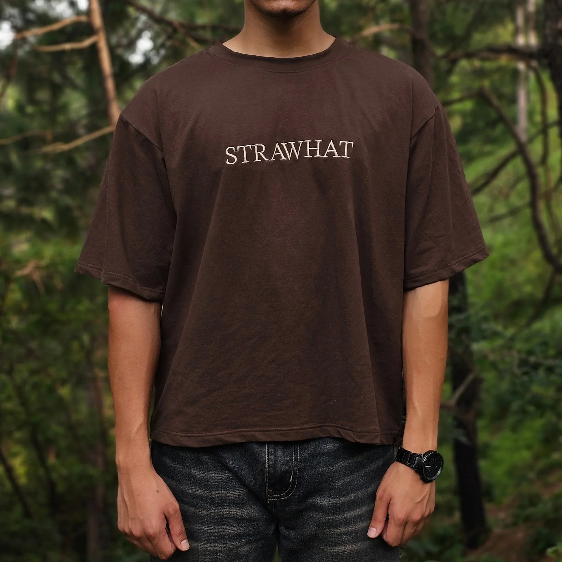 Person wearing a brown t-shirt with 'STRAWHAT' text in a forest setting