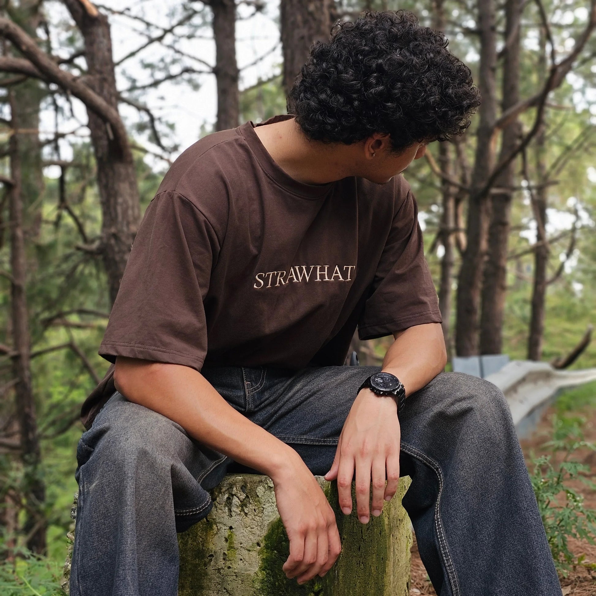 Person sitting on a log in a forest wearing a brown sweatshirt with 'STRAWHAT' branding.