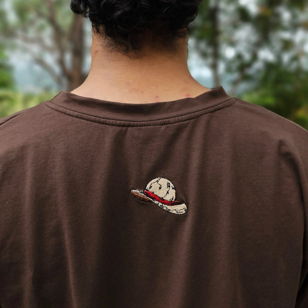 Brown shirt with a Anime Cap design on the Back embrioded against a blurred natural background