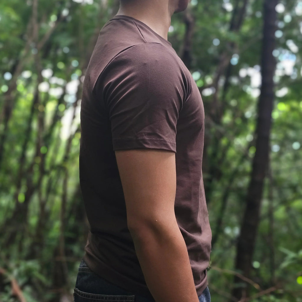 Person in a brown shirt and dark pants standing in a forest with green trees in the background