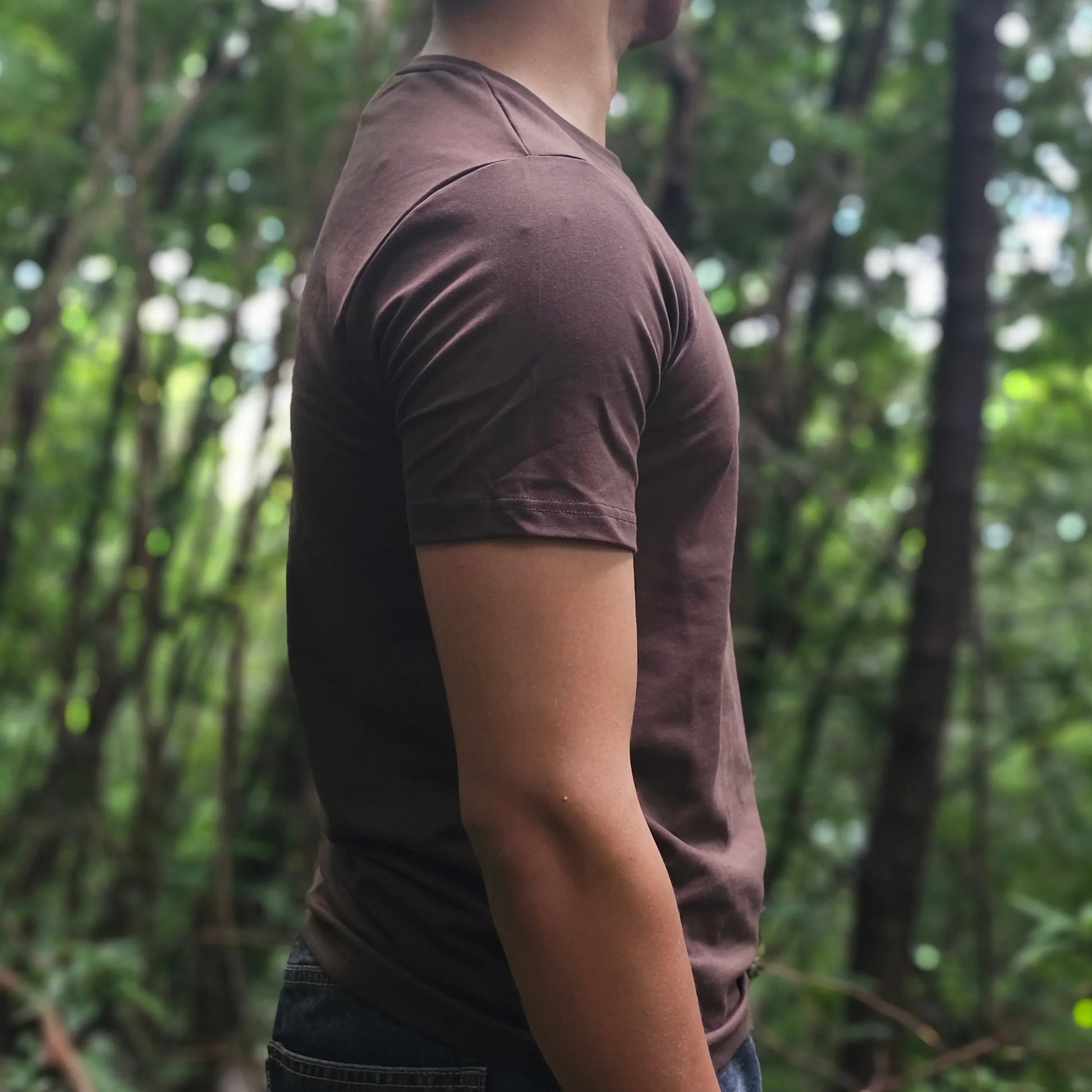Person in a brown shirt and dark pants standing in a forest with green trees in the background